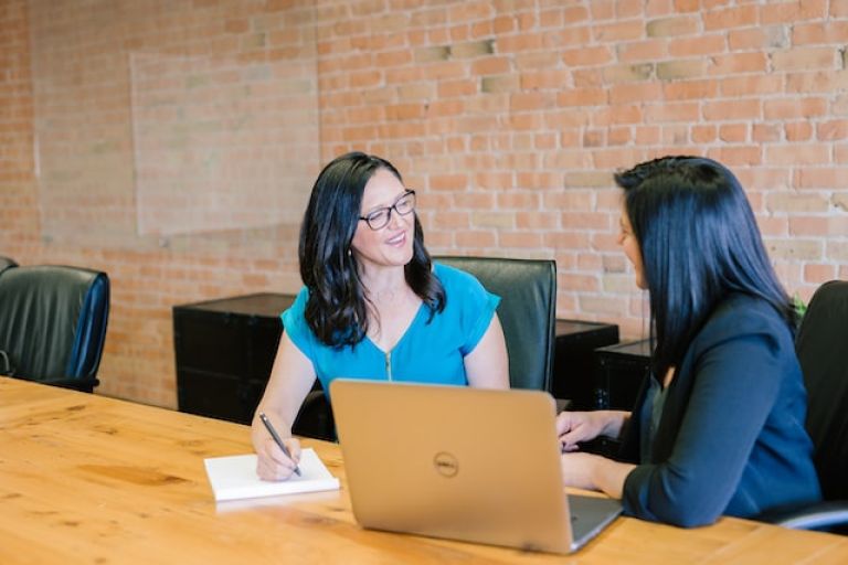 2 business women with a laptop and notepad discuss business in a conference room